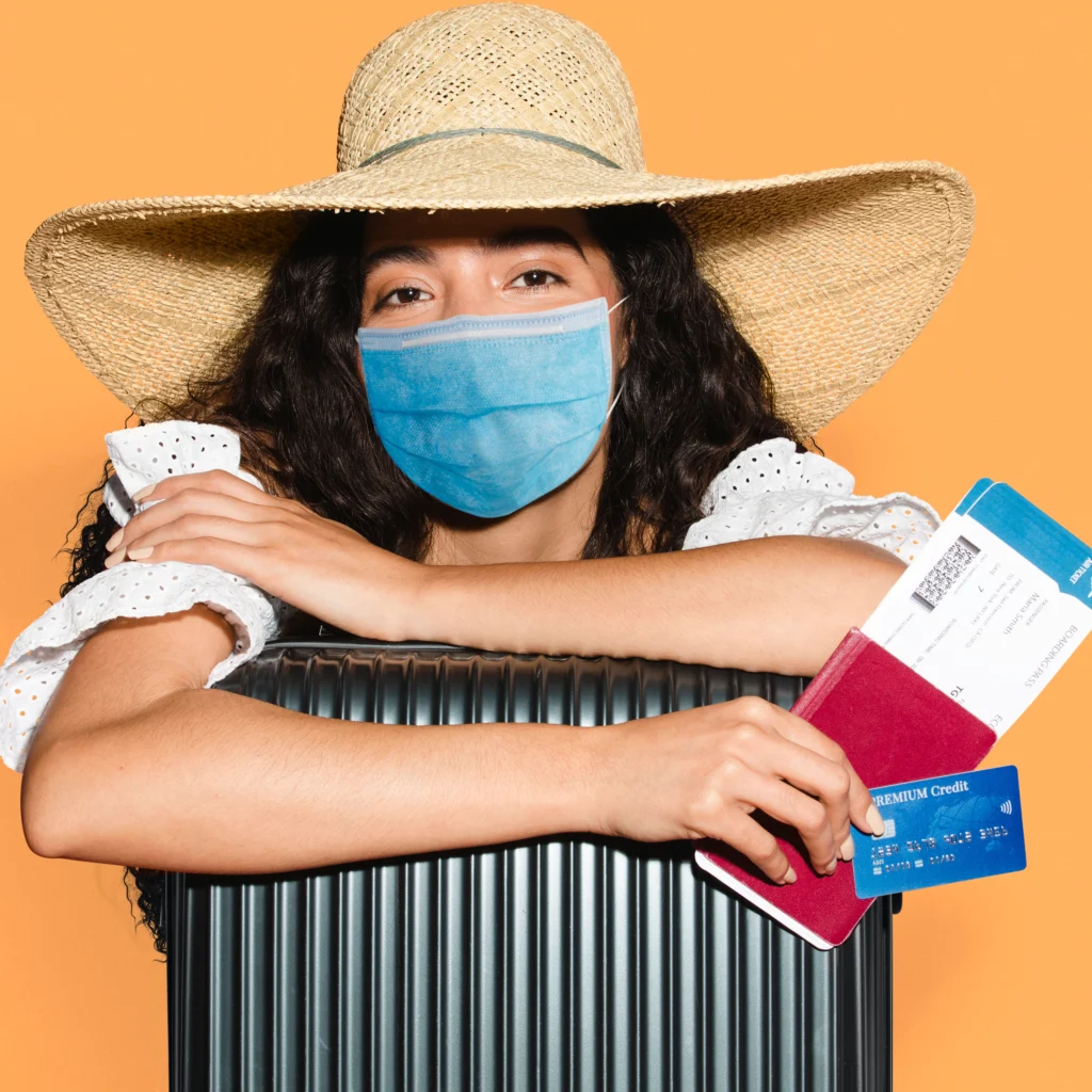 Woman wearing a face mask, straw hat, and holding travel documents and a credit card next to a suitcase, preparing for a trip.