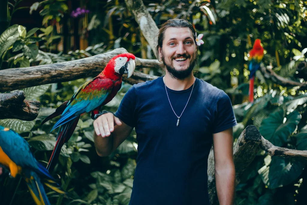 Smiling man with a flower in his hair, interacting with colorful macaws in a lush tropical setting.