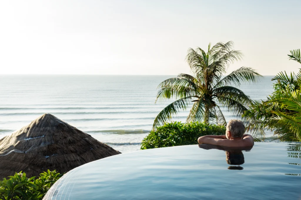 Man relaxing in a pool overlooking a calm ocean with palm trees and a thatched roof.