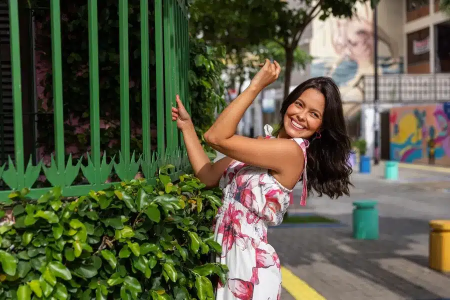 Smiling woman in a floral dress leans against a green metal fence in an urban setting.