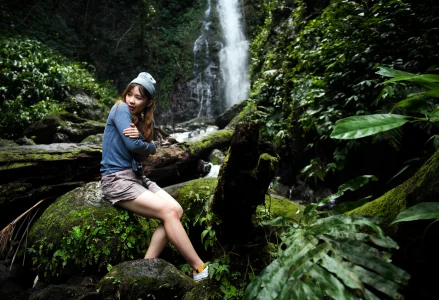 Woman wearing a beanie, sitting on a mossy rock by a waterfall in a lush rainforest.